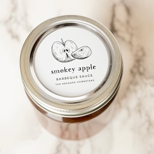 Może przedstawiać: Srebrny słoik z białą etykietą, na kt&oacute;rej widnieje napis "Smokey Apple Barbecue Sauce The Negaard Homestead". Etykieta zawiera r&oacute;wnież czarno-białą ilustrację jabłka.