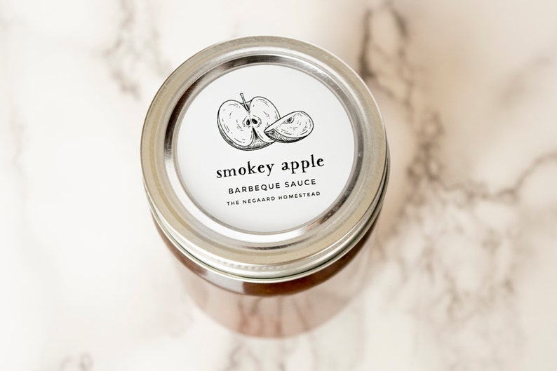 Może przedstawiać: Srebrny słoik z białą etykietą, na kt&oacute;rej widnieje napis "Smokey Apple Barbecue Sauce The Negaard Homestead". Etykieta zawiera r&oacute;wnież czarno-białą ilustrację jabłka.