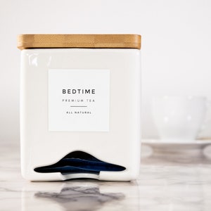 May include: White ceramic tea container with a bamboo lid. The container has a label that reads "BEDTIME PREMIUM TEA ALL NATURAL". The container has a slot for tea bags and a few tea bags are visible.