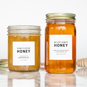 May include: Two glass jars of honey with labels. The jar on the left is labeled "Raw Clover Honey" and the jar on the right is labeled "Wildflower Honey".