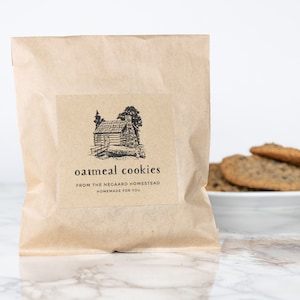 May include: A brown paper bag with a label that says "oatmeal cookies from the Negaard Homestead homemade for you". The label also features a black and white illustration of a cabin in the woods. A plate of oatmeal cookies is in the background.