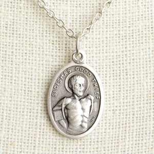 Może przedstawiać: Srebrny naszyjnik z zawieszką w postaci religijnego medalu z wizerunkiem Świętego Dismasza, "Dobrego Złodzieja", na przodzie. Medal ma owalny kształt i jest ozdobiony napisem "St. Dismas Good Thief" wokół krawędzi.