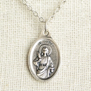 May include: Silver-toned oval pendant necklace featuring an image of St. Lucy. The pendant has the words "ST. LUCY" at the top and a depiction of a woman holding a palm branch and a small container. The necklace hangs on a delicate chain.
