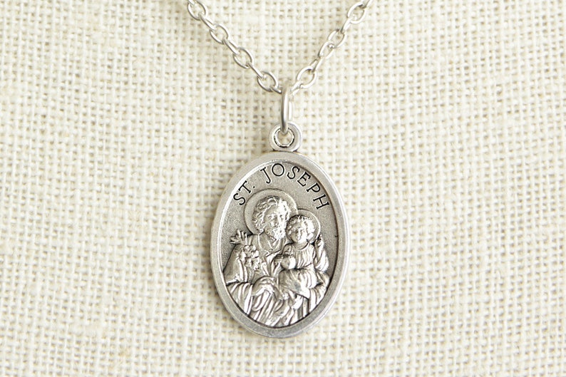 May include: Silver oval pendant necklace with a depiction of Saint Joseph holding baby Jesus. The pendant has the words "ST. JOSEPH" inscribed around the edge.