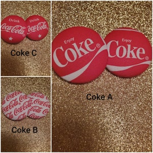 May include: Three different styles of Coca-Cola themed buttons. The first button has a red background with white Coca-Cola text and a white wave. The second button has a white background with red Coca-Cola text and a red wave. The third button has a white background with red Coca-Cola text repeated in a pattern.