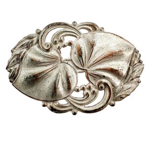 May include: A silver-toned brooch with a delicate design featuring two stylized leaves. The leaves are intricately detailed and have a slightly textured surface.