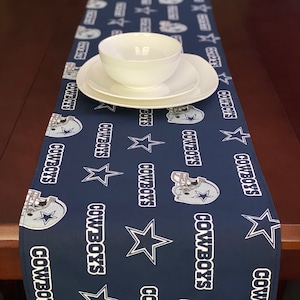 K&ouml;nnte beinhalten: Ein dunkelblauer Tischl&auml;ufer mit einem sich wiederholenden Muster aus wei&szlig;en Sternen, Footballhelmen und dem Wort "Cowboys".