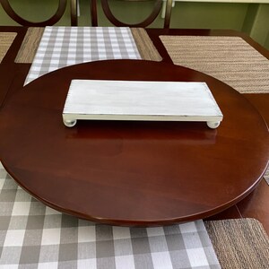 May include: A rectangular, white-painted wooden serving tray with rounded feet. The tray is placed on a large, dark brown, round table. A gray and white checkered runner is visible beneath the tray.