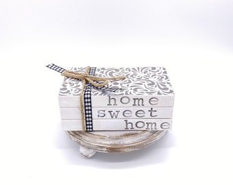 Op de afbeelding: Decoratieve houten blokkenset met de woorden "home sweet home" in zwarte letters. Het bovenste blok heeft een grijs en wit bloemenpatroon en de set is vastgebonden met een zwart-wit geruit lint en touw. De set staat op een kleine, ronde, witte sokkel.