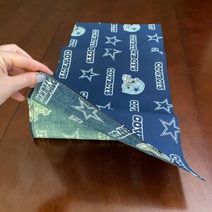 K&ouml;nnte beinhalten: Ein blauer Tischl&auml;ufer aus Stoff mit einem wei&szlig;en Stern und dem sich wiederholenden Muster "Dallas Cowboys". Der L&auml;ufer ist in der Mitte gefaltet und der Stoff ist strukturiert.