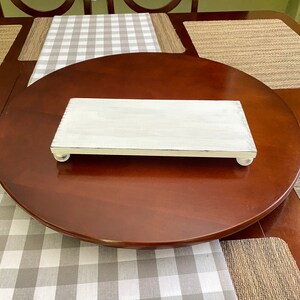 May include: A rectangular, white-painted wooden serving tray with rounded feet. The tray has a distressed finish and is placed on a dark brown, round table. A gray and white checkered table runner is visible.