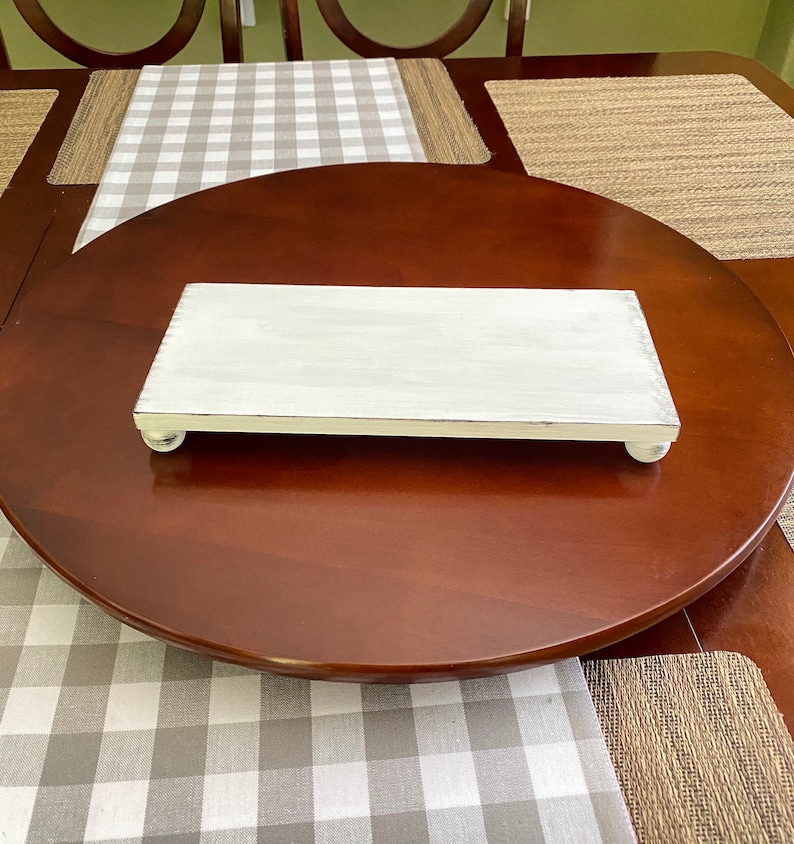 May include: A rectangular, white-painted wooden serving tray with rounded feet. The tray has a distressed finish and is placed on a dark brown, round table. A gray and white checkered table runner is visible.