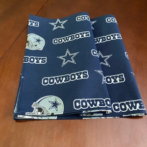 K&ouml;nnte beinhalten: Zwei dunkelblaue Stoff-Platzsets mit einem wei&szlig;en Stern und dem Wort "COWBOYS", das darauf wiederholt wird. Die Platzsets zeigen auch einen wei&szlig;en Umriss eines Footballhelms mit einem Stern an der Seite.