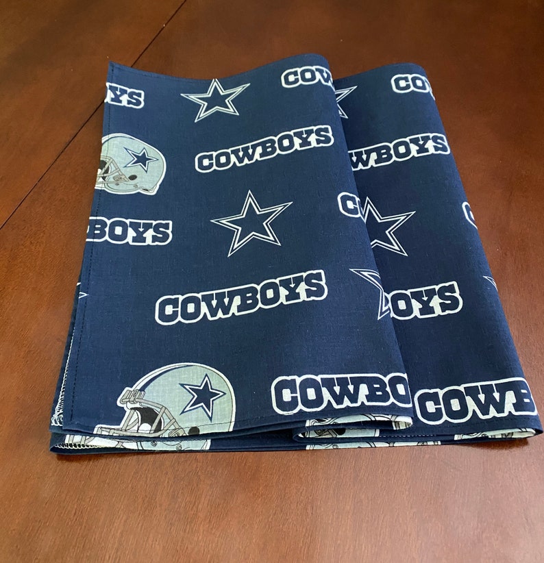 K&ouml;nnte beinhalten: Zwei dunkelblaue Stoff-Platzsets mit einem wei&szlig;en Stern und dem Wort "COWBOYS", das darauf wiederholt wird. Die Platzsets zeigen auch einen wei&szlig;en Umriss eines Footballhelms mit einem Stern an der Seite.