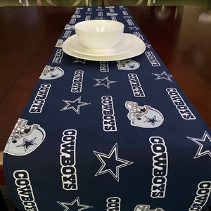 K&ouml;nnte beinhalten: Ein marineblauer Tischl&auml;ufer mit einem sich wiederholenden Muster aus wei&szlig;en Sternen, dem Wort "Cowboys" und einem Footballhelm. Der L&auml;ufer liegt auf einem Holztisch mit einer wei&szlig;en Sch&uuml;ssel und einem Teller darauf.