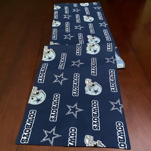 K&ouml;nnte beinhalten: Ein dunkelblauer Tischl&auml;ufer mit einem sich wiederholenden Muster aus wei&szlig;en Sternen, Footballhelmen und den Worten "Cowboys".