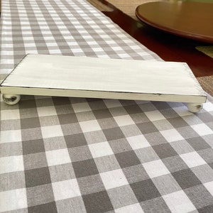 May include: A rectangular, white-painted wooden riser with distressed edges and ball feet. The riser is displayed on a gray and white checkered table runner, suitable for displaying decorative items.