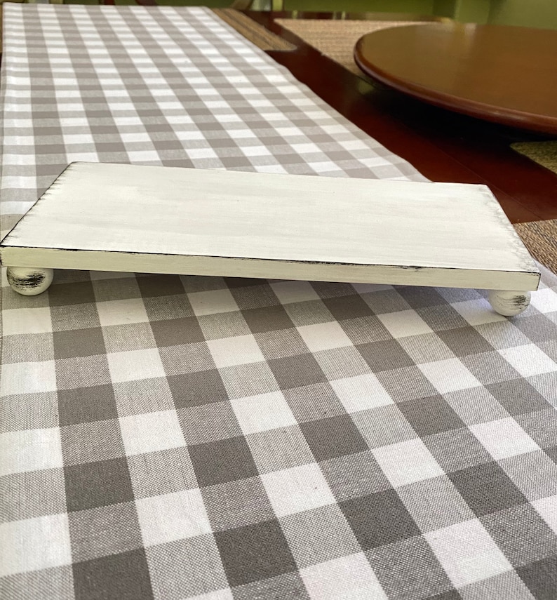 May include: A rectangular, white-painted wooden riser with distressed edges and ball feet. The riser is displayed on a gray and white checkered table runner, suitable for displaying decorative items.