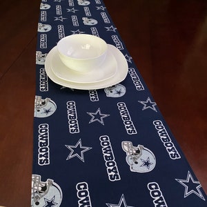 K&ouml;nnte beinhalten: Ein dunkelblauer Tischl&auml;ufer mit einem Muster aus wei&szlig;en Sternen und Footballhelmen. Das Muster wiederholt die Worte "Cowboys" durchgehend. Eine wei&szlig;e Sch&uuml;ssel und ein Teller liegen auf dem L&auml;ufer.