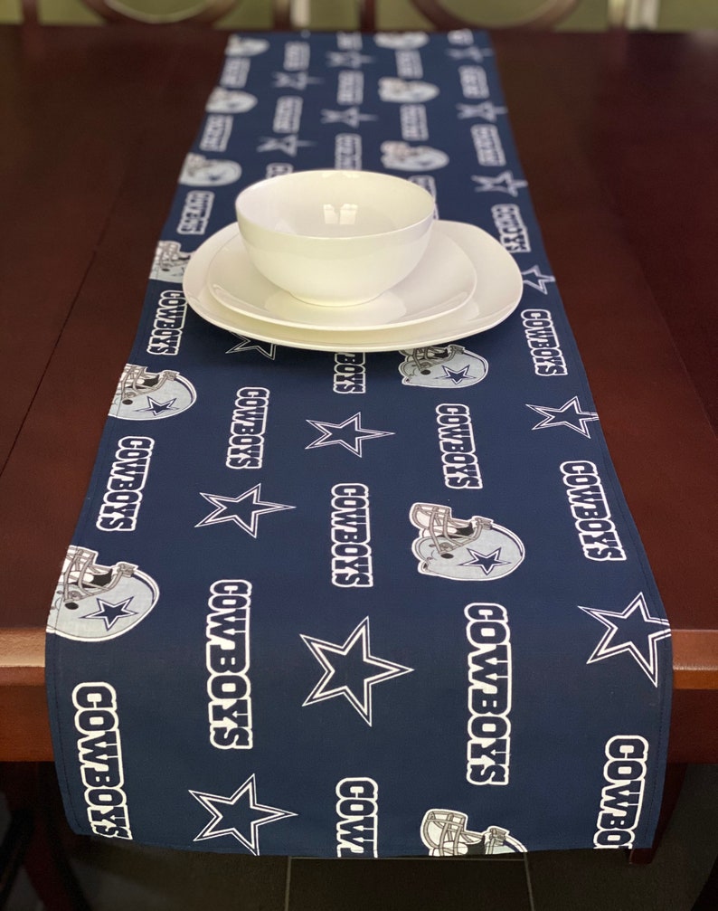 K&ouml;nnte beinhalten: Ein dunkelblauer Tischl&auml;ufer mit einem sich wiederholenden Muster aus wei&szlig;en Sternen, Footballhelmen und dem Wort "Cowboys".