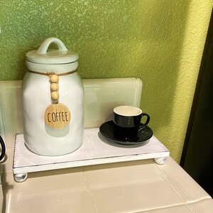 May include: A white wooden riser with a white ceramic coffee canister and a black coffee cup and saucer. The canister has a lid and a wooden tag that says "COFFEE". The riser has small, round feet.