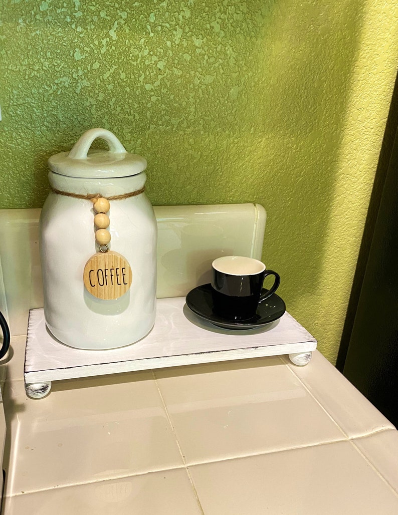 May include: A white wooden riser with a white ceramic coffee canister and a black coffee cup and saucer. The canister has a lid and a wooden tag that says "COFFEE". The riser has small, round feet.