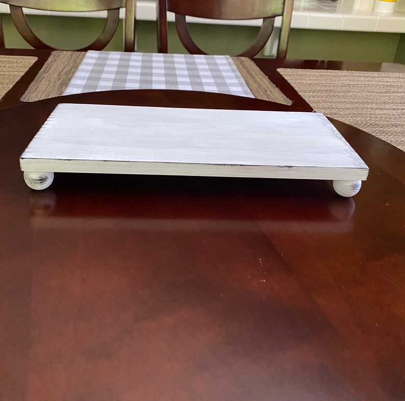 May include: A rectangular, white-painted wooden serving tray with rounded feet. The tray has a distressed finish, with visible wood grain and slight imperfections. The tray is designed for displaying food or decorative items.