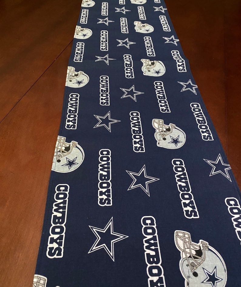 K&ouml;nnte beinhalten: Ein blauer Tischl&auml;ufer mit einem sich wiederholenden Muster aus wei&szlig;en Sternen, den Worten "COWBOYS" und einem Footballhelm.