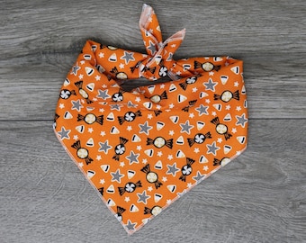 Puede incluir: Bandana naranja con un patrón de dulces de Halloween. La bandana presenta maíz dulce negro y blanco, piruletas y estrellas.
