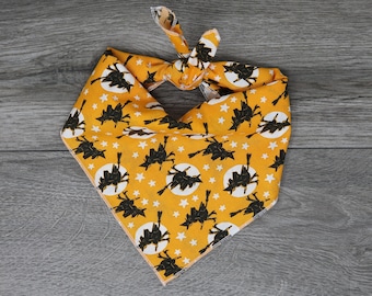 Puede incluir: Un pañuelo amarillo con un estampado en blanco y negro de brujas volando en escobas y estrellas. El pañuelo está atado en un nudo.
