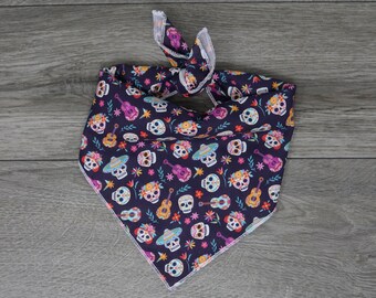 Puede incluir: Un pañuelo de color morado oscuro con ribete blanco. El pañuelo presenta un patrón repetido de calaveras de azúcar coloridas, flores y guitarras.
