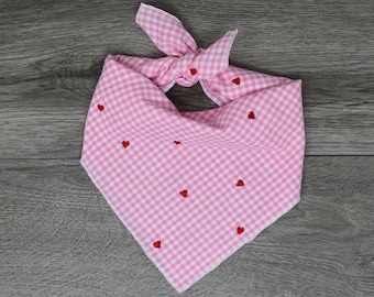 Könnte beinhalten: Rosa-weißes Vichy-Hundebandana mit roten Herzstickereien. Das Bandana ist oben verknotet und liegt auf einer grauen Holzoberfläche. Es hat eine dreieckige Form.