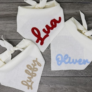Pode incluir: Três bandanas brancas para cães com nomes bordados em fio vermelho, bege e azul. Os nomes "Lua", "Luffy" e "Oliver" são exibidos nas bandanas, que são amarradas no topo. As bandanas são feitas de um tecido macio e texturizado.