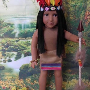 May include: A doll dressed in Native American attire, featuring a headdress with colorful feathers and a beaded headband. The doll wears a tan dress with a striped hem and holds a spear. Small toy chicks and a pouch with stones are at the doll's feet.