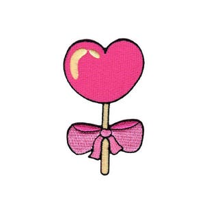 Puede incluir: Parche bordado con un caramelo en forma de corazón rosa con un resaltado amarillo y un lazo rosa. El palo del caramelo es marrón y el diseño está delineado en negro. Sobre un fondo blanco.