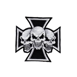 May include: A black iron cross with three white skulls in the centre. The skulls are facing forward with their eyes open and their mouths slightly open.