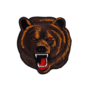 Op de afbeelding: Een close-up van het gezicht van een bruine beer met zijn bek open, waardoor scherpe tanden zichtbaar zijn. De beer heeft oranje ogen en een donkerbruine neus.