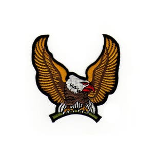 Könnte beinhalten: Ein gestickter Aufnäher mit einem braunen und weißen Adler, dessen Flügel weit ausgebreitet sind. Der Adler hat einen roten Schnabel und gelbe Krallen. Der Aufnäher befindet sich auf einem weißen Hintergrund.