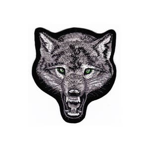 Op de afbeelding: Gestikte patch van een wolfskop met een grommende uitdrukking en groene ogen. De wolf is grijs en de achtergrond is zwart.