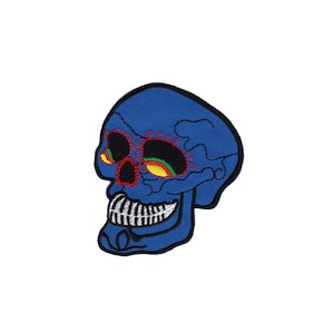Könnte beinhalten: Blauer gestickter Totenkopf-Patch mit roten und gelben Details. Der Schädel hat ein zahniges Grinsen und komplizierte Augenmuster.