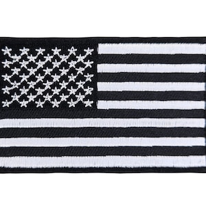Könnte beinhalten: Schwarzer und weißer gestickter amerikanischer Flaggen-Patch. Der Patch zeigt 13 weiße Streifen und 50 weiße Sterne auf schwarzem Hintergrund.