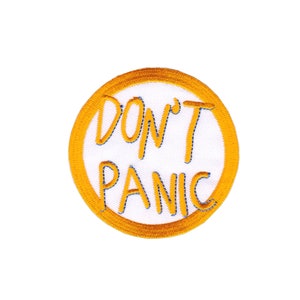 Könnte beinhalten: Ein weißer gestickter Aufnäher mit einem gelben Kreisrand. Der Text "DON'T PANIC" ist in Gelb gestickt.
