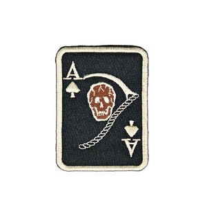 Könnte beinhalten: Ein gestickter Patch in Schwarz und Weiß, der ein As Pik mit einem Totenkopf und einer Sense zeigt. Die Karte ist weiß umrandet.