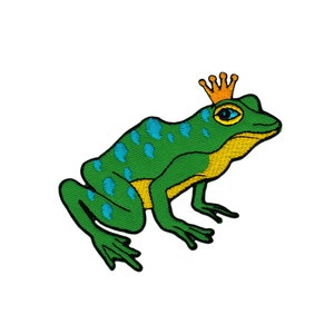 Op de afbeelding: Een groene kikker met blauwe vlekken die een gouden kroon draagt. De kikker zit op een witte achtergrond.