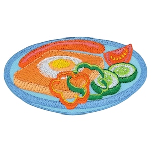 Könnte beinhalten: Ein gestickter Bügel-Patch, der einen Teller mit Essen zeigt. Der Teller ist blau und zeigt eine Scheibe Brot mit einem Ei, einer Wurst, einem roten Paprika und Gurkenscheiben.