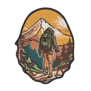 Könnte beinhalten: Gestickter Aufnäher mit einem Wanderer mit Rucksack, der auf eine Bergkette zugeht. Der Aufnäher hat einen gelb-orangen Sonnenuntergangshintergrund mit Bäumen und einem Pfad im Vordergrund. Der Wanderer trägt eine grüne Jacke und eine orangefarbene Hose.