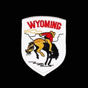 Könnte beinhalten: Weißer gestickter Aufnäher mit dem Wort "WYOMING" in roten Buchstaben. In der Mitte des Aufnähers befindet sich ein Cartoon-Bild eines Cowboys, der ein buckelndes Pferd reitet.