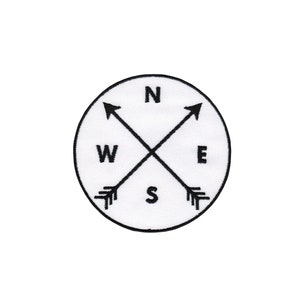 Könnte beinhalten: Weißer runder Patch mit schwarzen gestickten Pfeilen, die in die Richtungen Nord, Süd, Ost und West zeigen. Die Buchstaben N, S, E und W sind ebenfalls in Schwarz gestickt.
