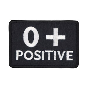 Op de afbeelding: Zwarte geborduurde patch met witte tekst die "O+ POSITIEF" luidt.