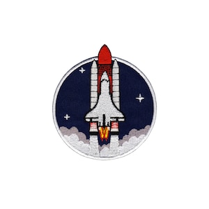 Op de afbeelding: Geborduurde patch met een spaceshuttle die gelanceerd wordt. Het ontwerp toont een witte shuttle, een rode booster en een donkerblauwe achtergrond met sterren. De patch is cirkelvormig met een witte rand.